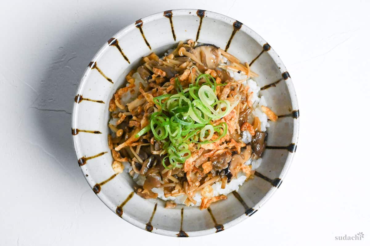 Fried mushrooms over rice sprinkled with crispy onion bits and chopped green onions in a white bowl with brown stripes