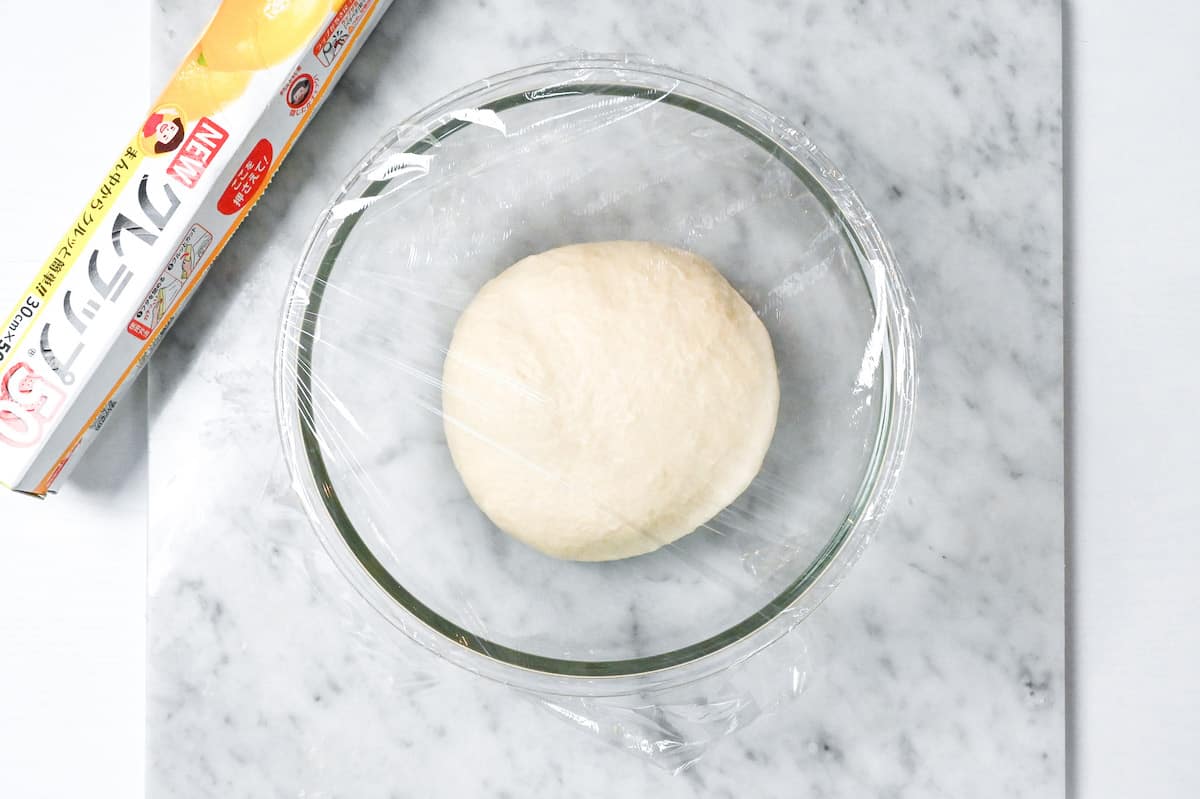 nama shokupan dough resting in a glass bowl covered with plastic wrap