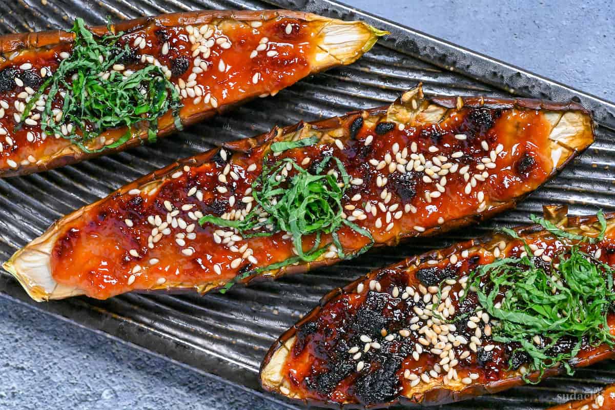 Nasu dengaku (Japanese miso-glazed eggplant) sprinkled with sesame seeds and shredded Perilla leaves on a dark textured rectangular plate on a gray background