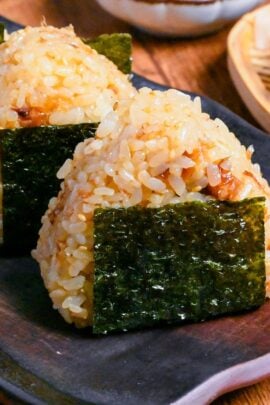 3 okaka onigiri (seasoned bonito flake rice balls) lined up on a dark brown rectangular plate on a wooden background