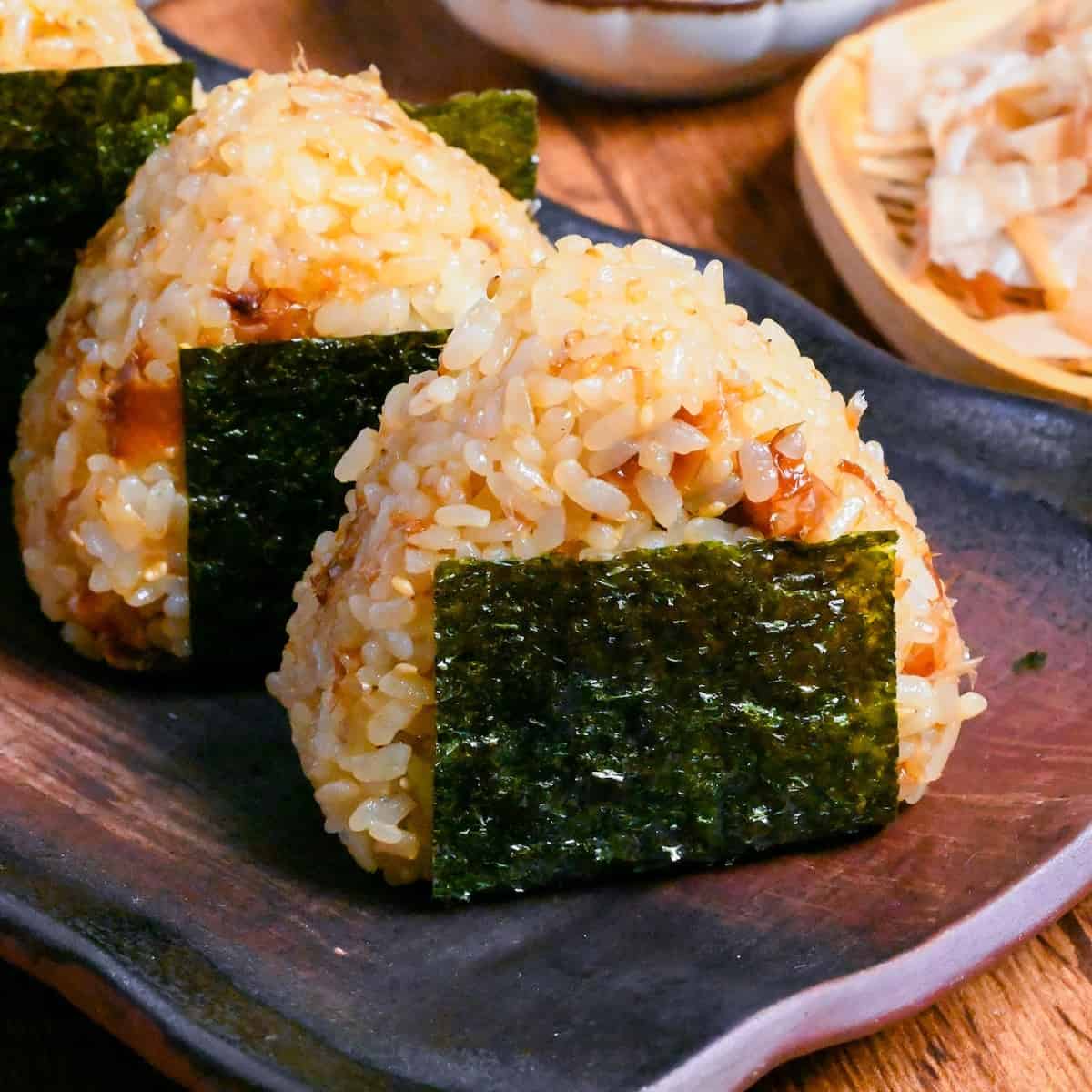 3 okaka onigiri (seasoned bonito flake rice balls) lined up on a dark brown rectangular plate on a wooden background