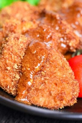 Oven-baked chicken katsu pieces placed over salad leaves and drizzled with sesame sauce on a black plate close up thumbnail