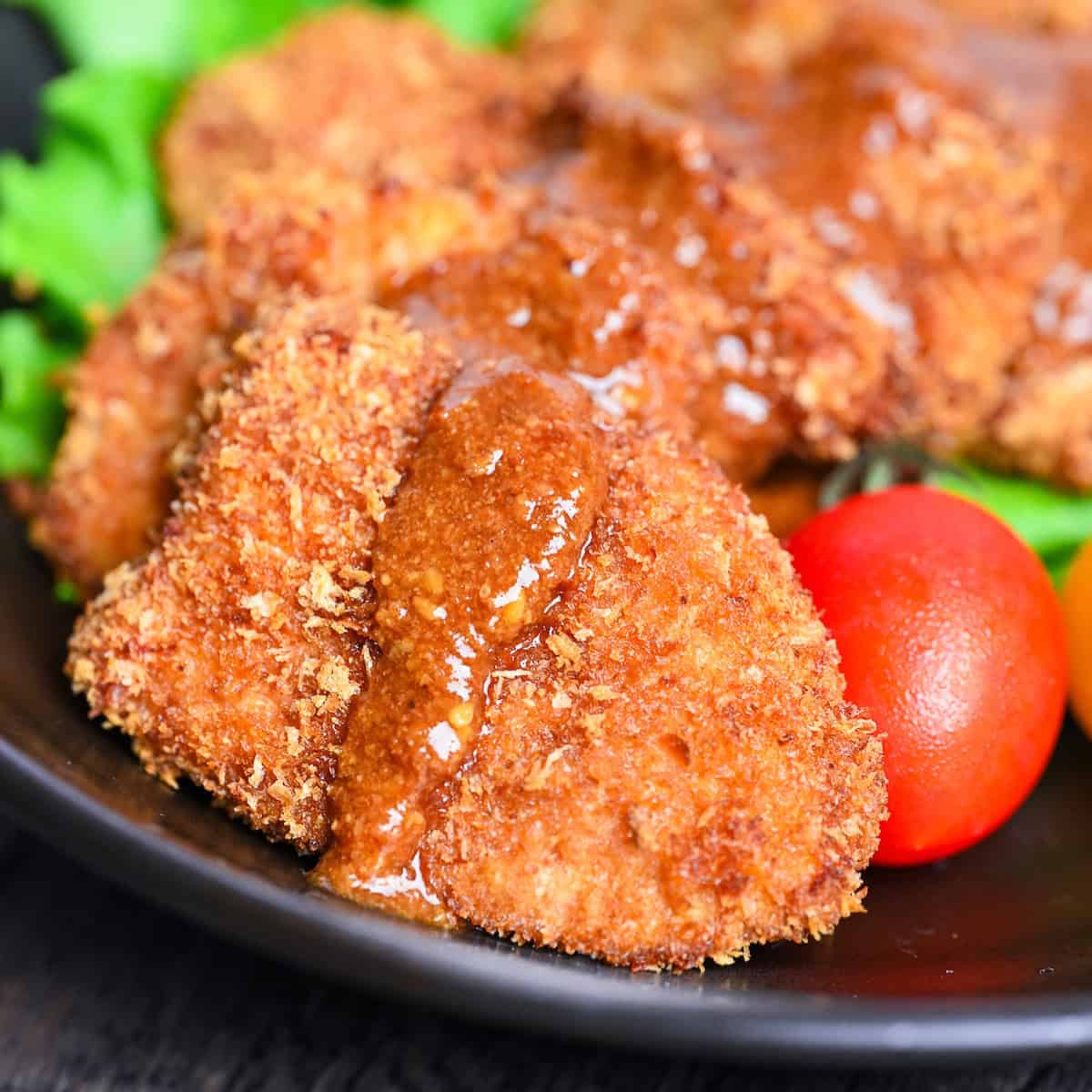 Oven-baked chicken katsu pieces placed over salad leaves and drizzled with sesame sauce on a black plate close up thumbnail