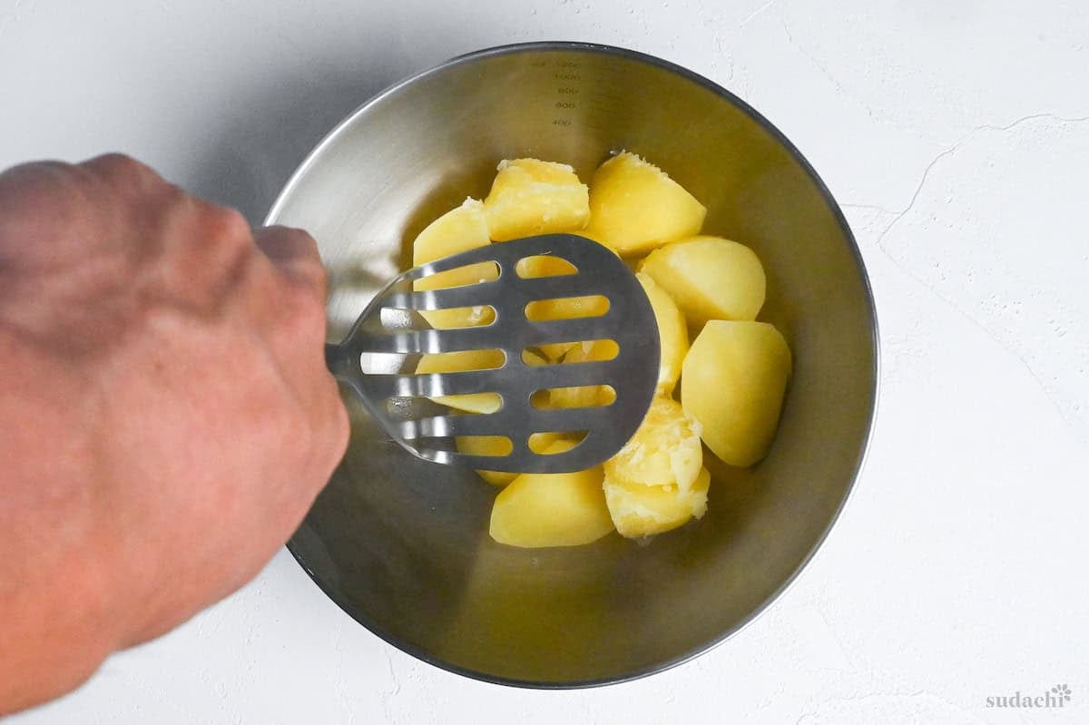 cooked potatoes in a mixing bowl being mashed with a potato masher