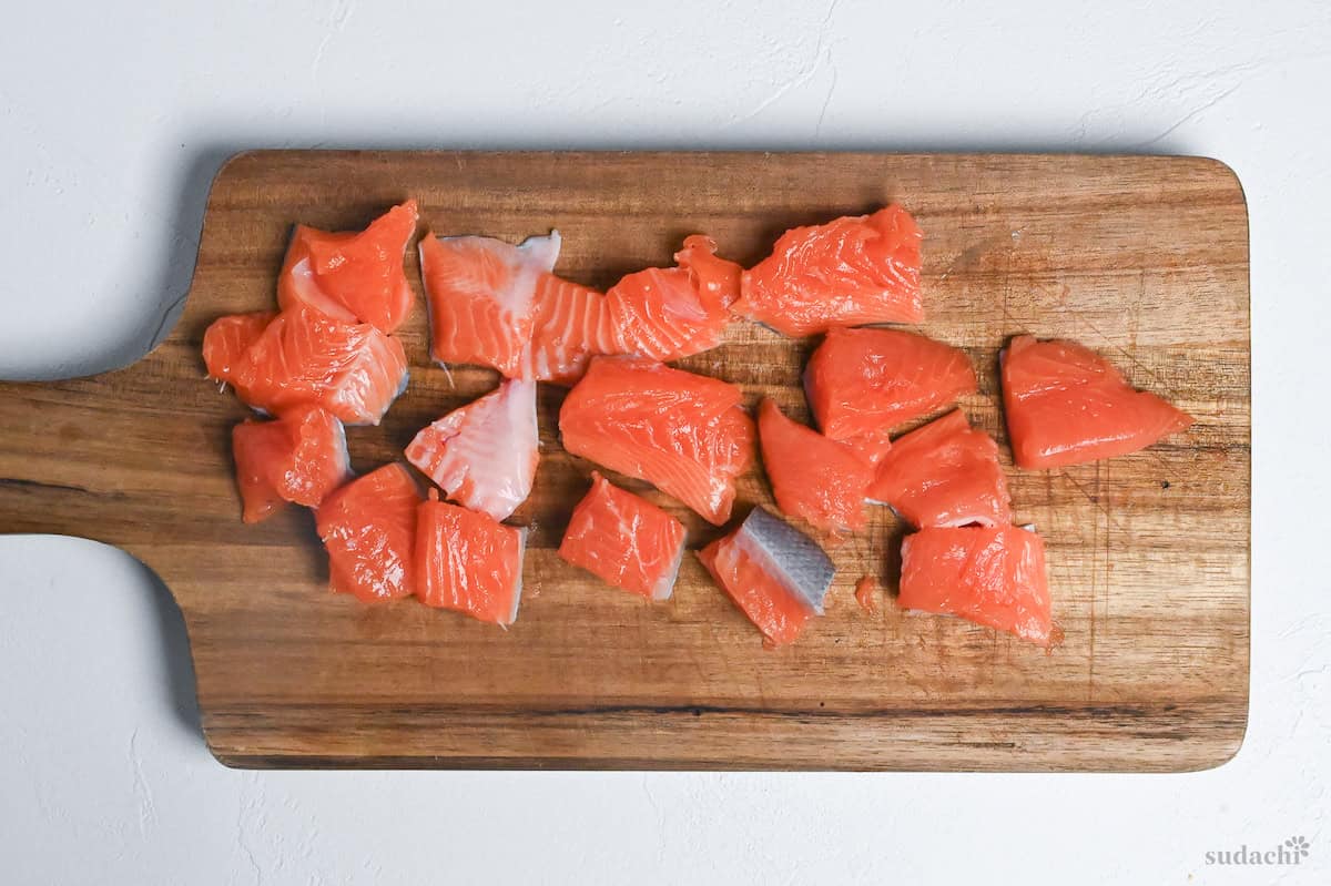 salmon fillet cut into bitesize pieces on a wooden chopping board on a white background