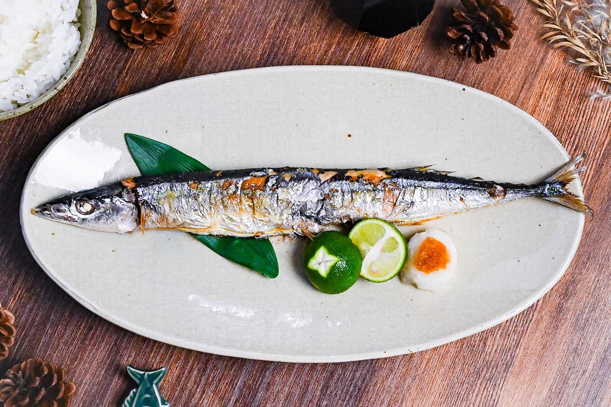 Grilled pacific saury (sanma no shioyaki) on a pale oval plate with grated daikon, sauce sauce and a halved sudachi
