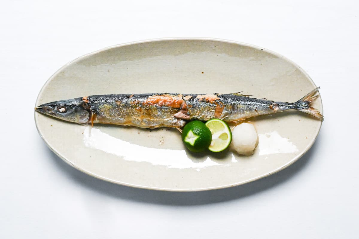 Grilled pacific saury (sanma no shioyaki) on a pale oval plate with grated daikon and a halved sudachi