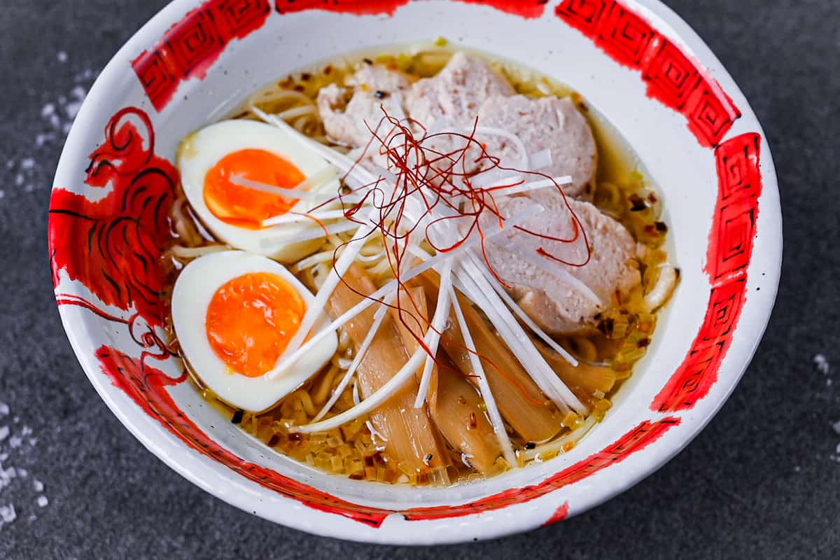 Japanese shio ramen topped with slices of chicken, menma (bamboo shoots), soft boiled egg, white spring onion and chilli threads in a red and white ramen bowl on a grey background