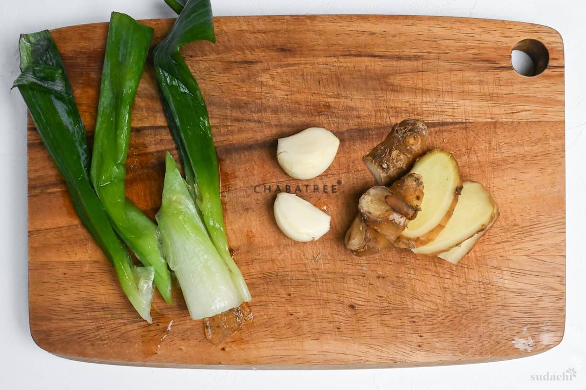 Green part of Japanese leek, 2 garlic cloves and sliced ginger root on a wooden cutting board