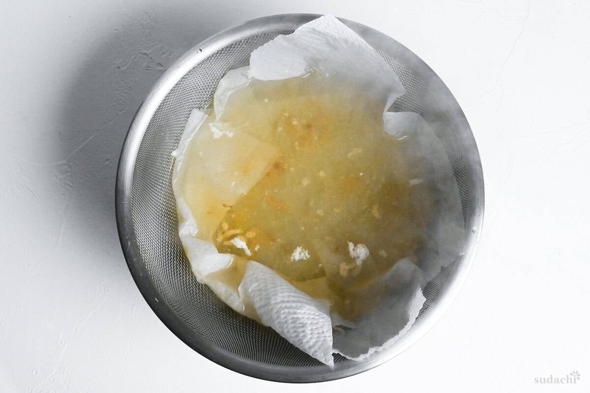 straining homemade shio ramen broth through kitchen paper over a sieve lined bowl