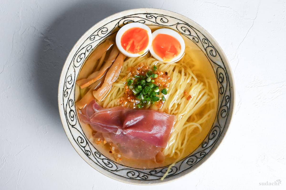 homemade shio ramen from scratch topped with soft boiled eggs, cured ham, menma bamboo shoots, crispy fried leeks and chopped green onion.