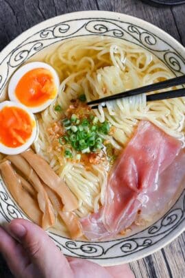 Homemade shio ramen topped with soft boiled eggs, menma, and ham in a cream bowl with black patterned rim on ashy wood background