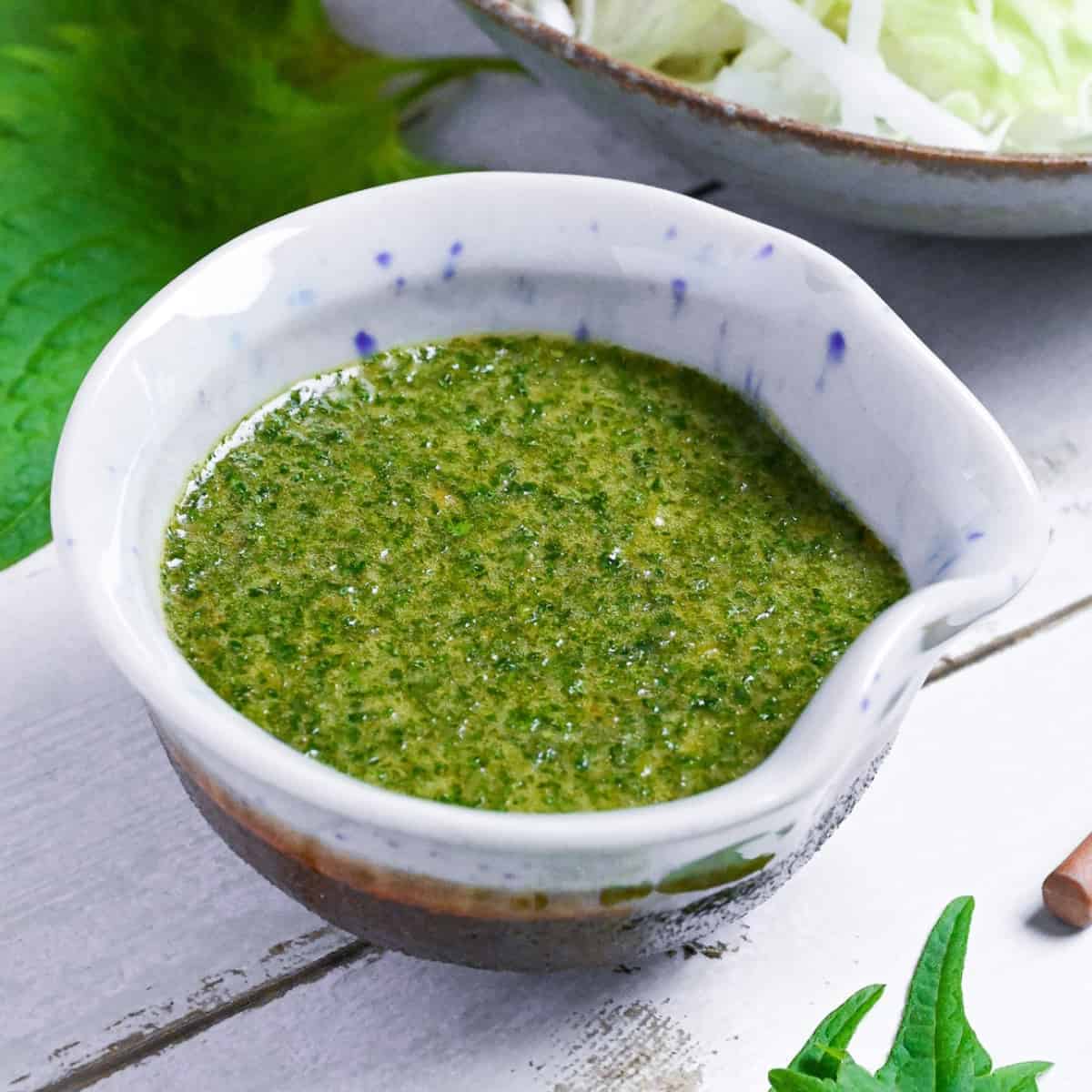 Japanese shiso dressing in a small blue and brown ceramic jug with a bowl of salad in the background and shiso leaves scattered around