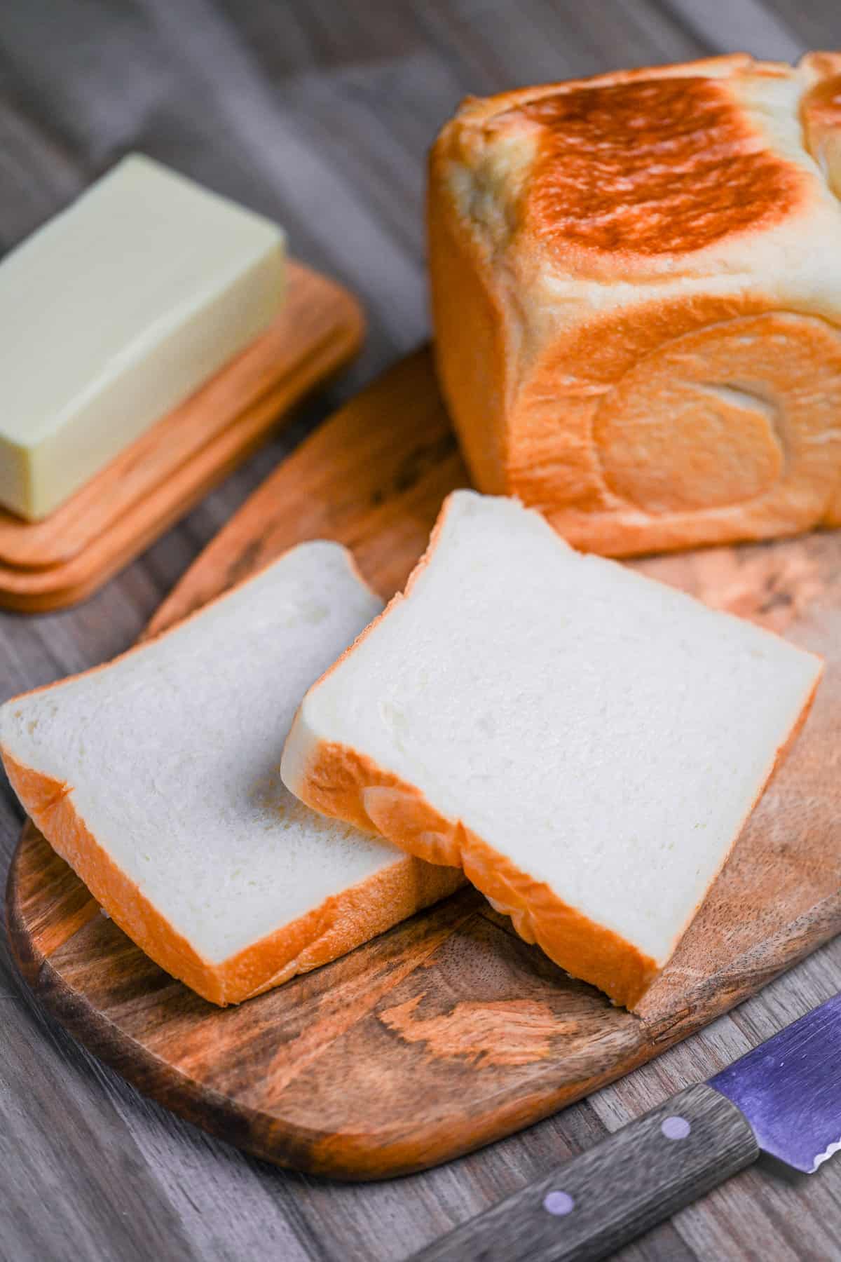 two slices of nama shokupan on a wooden chopping board with a block of butter and whole loaf in the background
