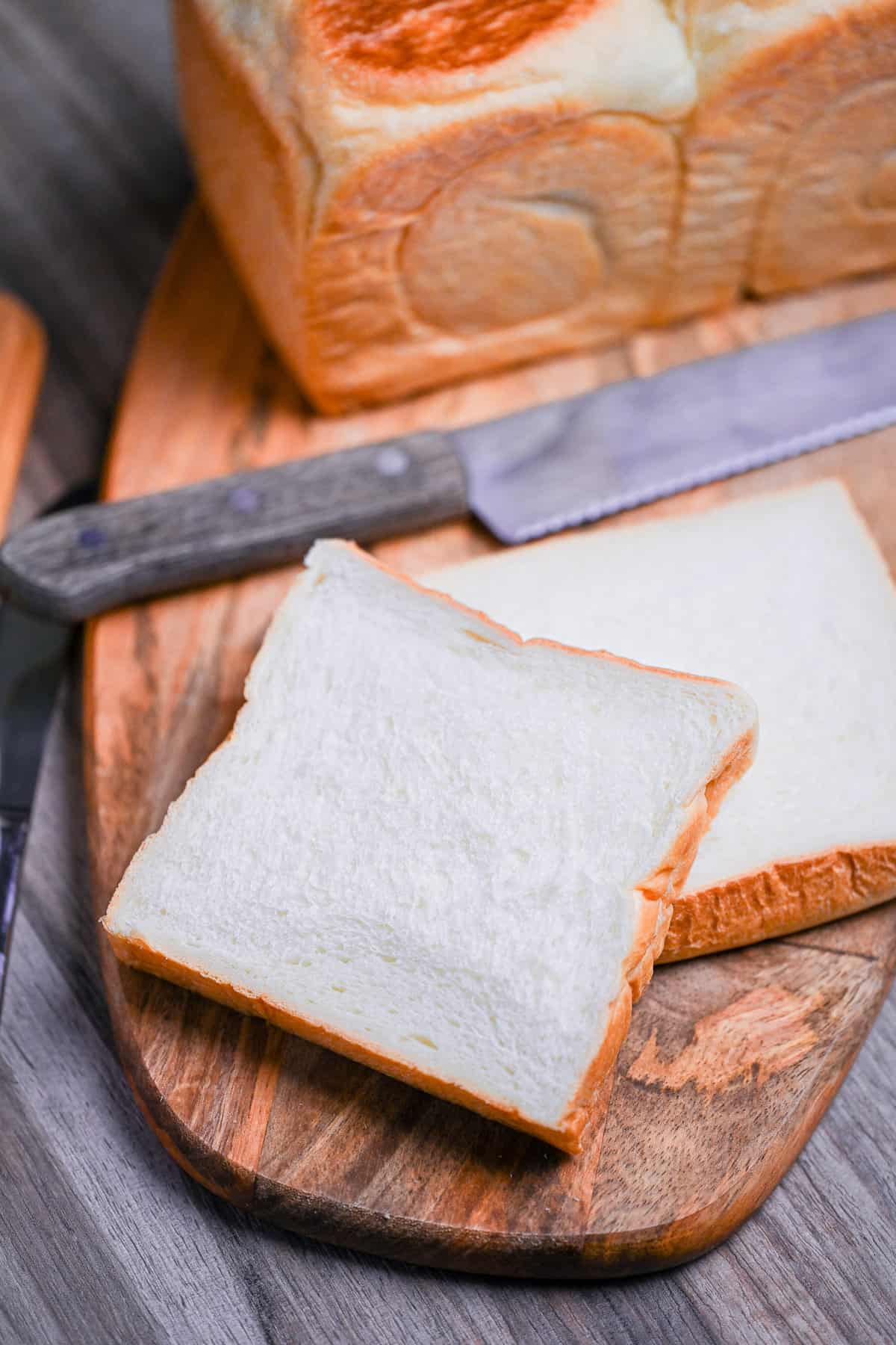 two slices of nama shokupan on a wooden chopping board with a bread knife and whole loaf in the background
