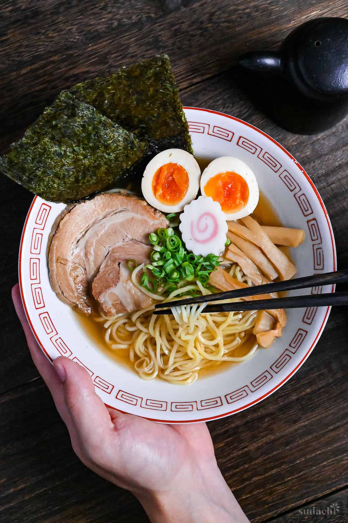 Homemade shoyu ramen in a white and red ramen bowl topped with pork chashu, nori, ramen eggs, narutomaki, chopped green onions and menma with noodles lifted with black chopsticks