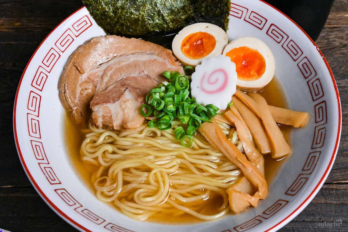 Homemade shoyu ramen in a white and red ramen bowl topped with pork chashu, ramen eggs, narutomaki, chopped green onions and menma on a dark wooden background close up