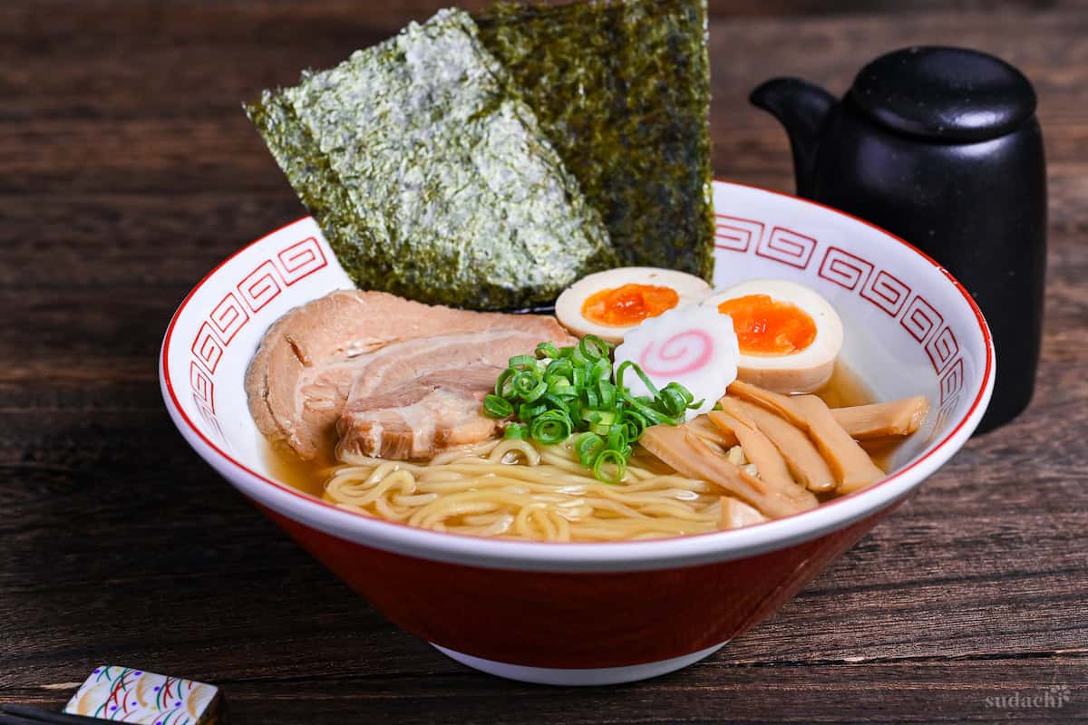Homemade shoyu ramen in a white and red ramen bowl topped with pork chashu, ramen eggs, narutomaki, chopped green onions and menma on a dark wooden background with black soy sauce bottle in the background side view