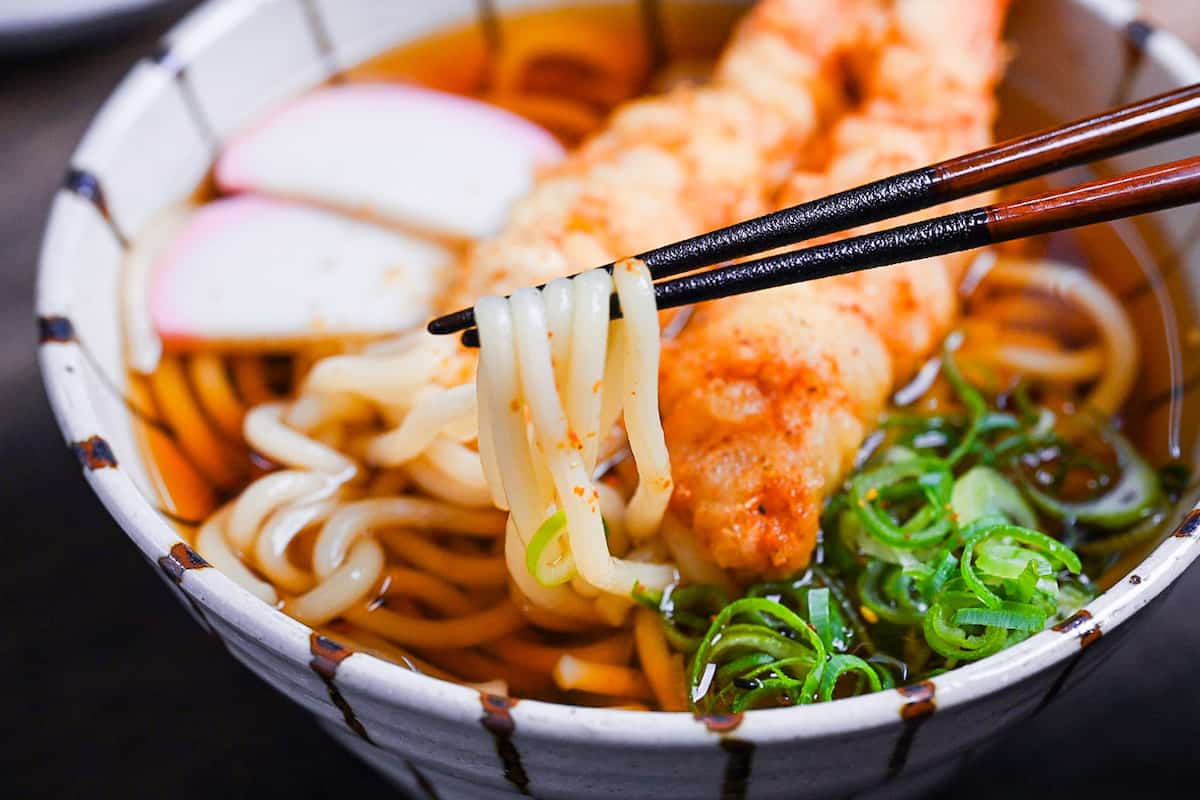 Ebiten Udon - Homemade udon noodle soup in a stripy bowl topped with kamaboko, green onion and tempura shrimp