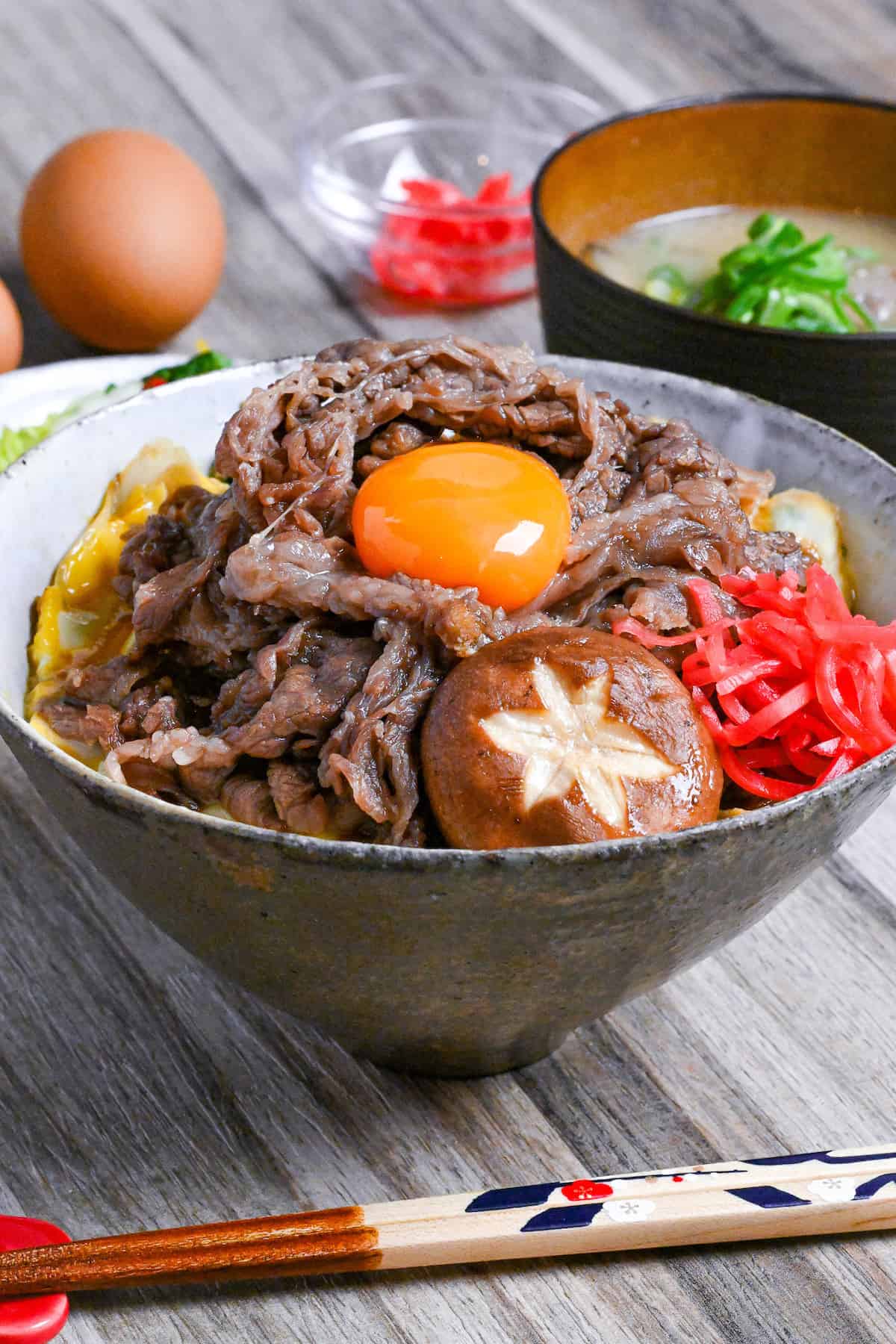 Sukiyaki beef donburi rice bowl topped with red pickled ginger, shiitake mushroom and egg yolk in a mottled bowl