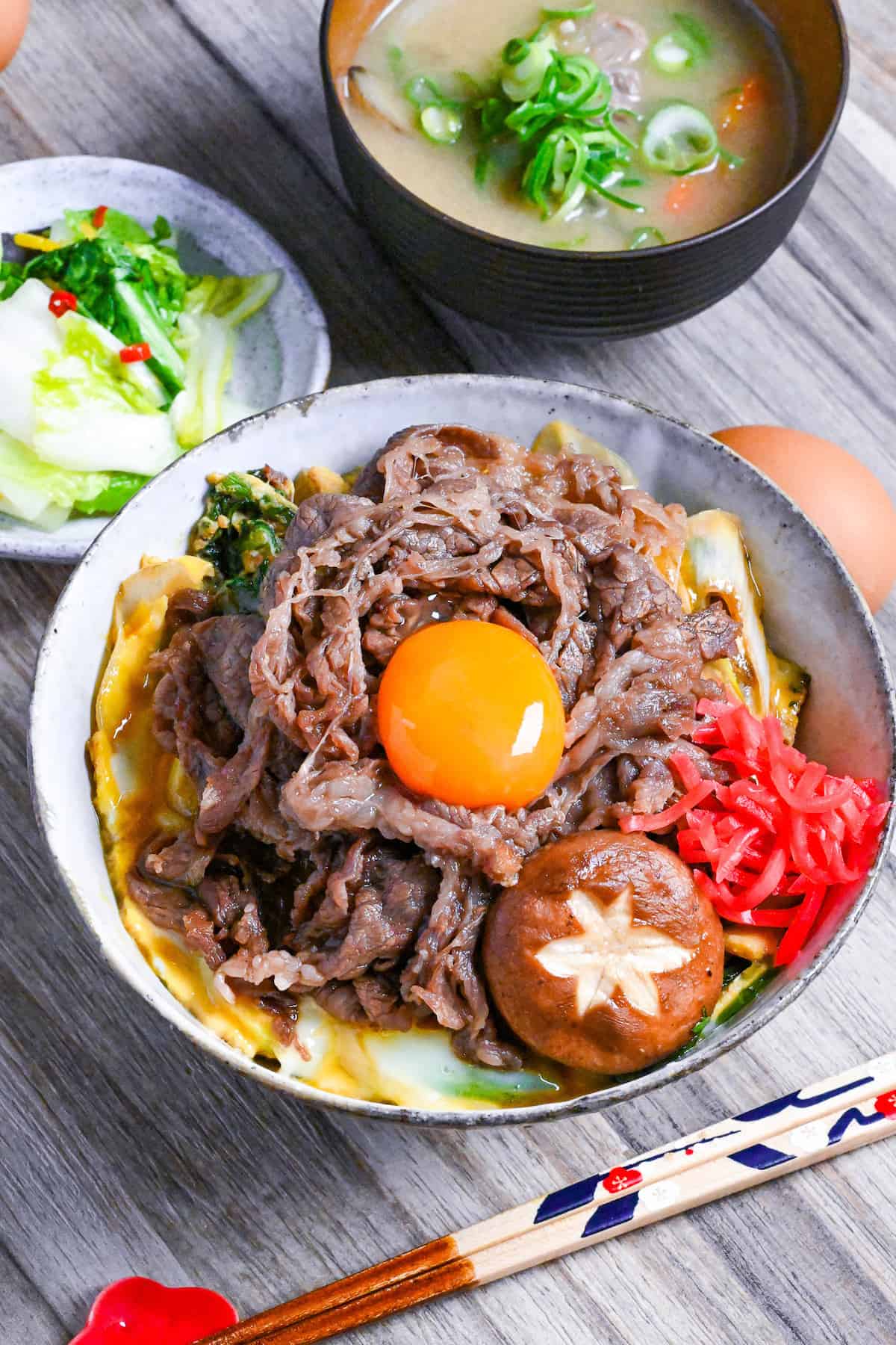 Sukiyaki beef donburi rice bowl topped with red pickled ginger, shiitake mushroom and egg yolk in a mottled bowl