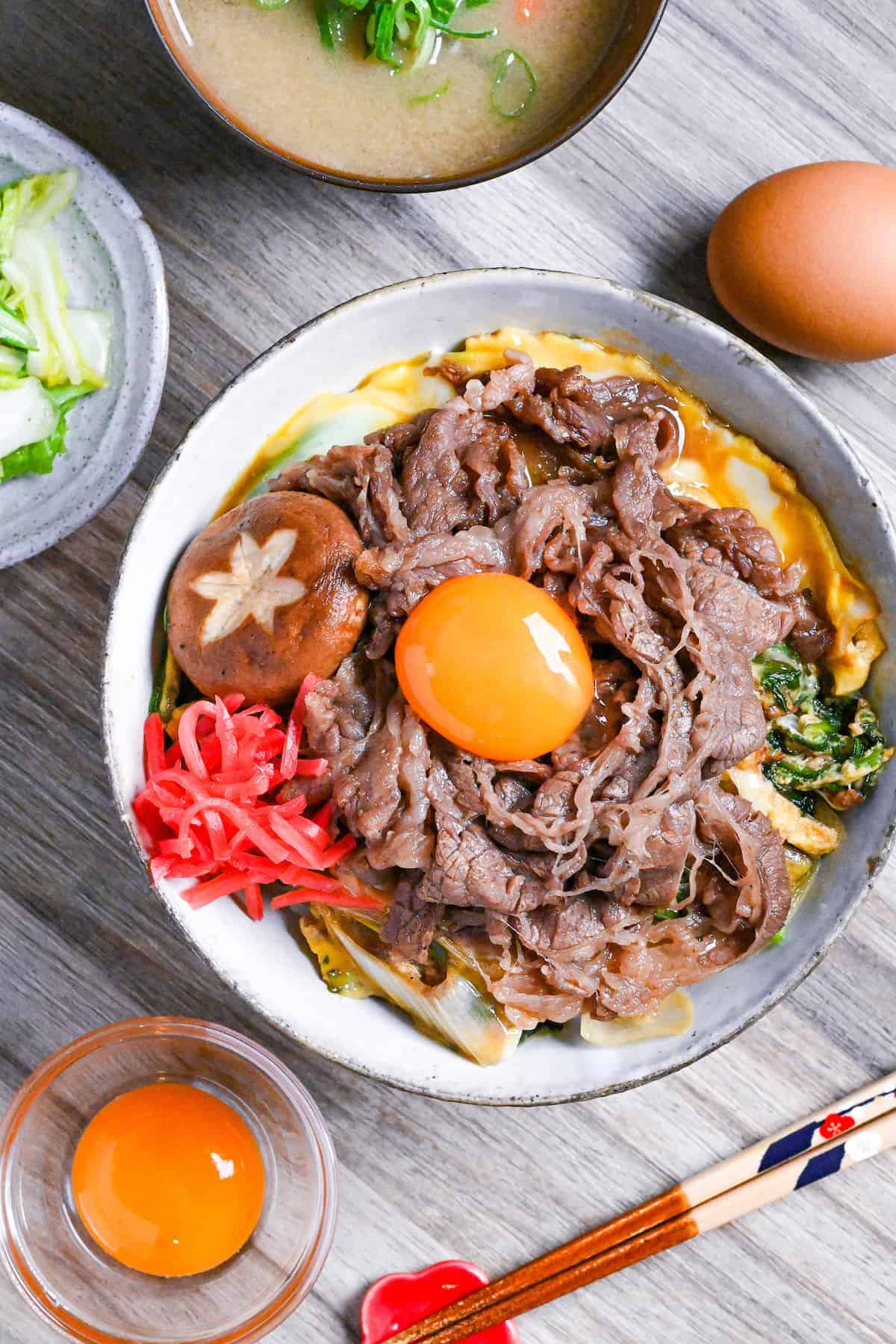 Sukiyaki beef donburi rice bowl topped with red pickled ginger, shiitake mushroom and egg yolk in a mottled bowl top down