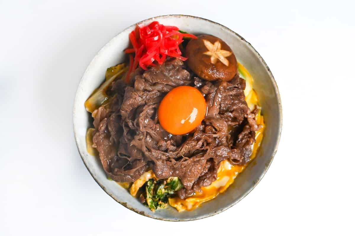 Sukiyaki beef donburi rice bowl topped with red pickled ginger, shiitake mushroom and egg yolk in a mottled bowl