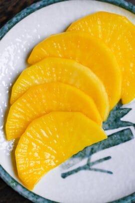 5 pieces of yellow pickled daikon radish (takuan) cut into half moons and arranged in a row on a white plate with green ink design close up