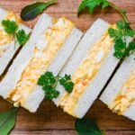 Four Japanese egg finger sandwiches (tamago sando) on a wooden chopping board with salad leaves