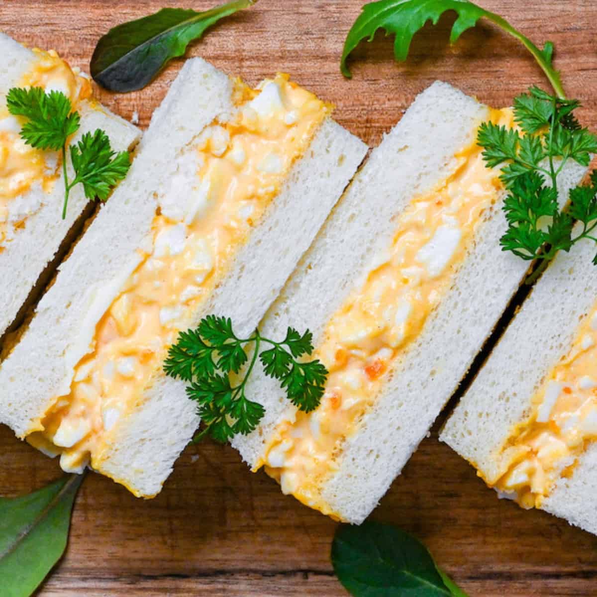 Four Japanese egg finger sandwiches (tamago sando) on a wooden chopping board with salad leaves
