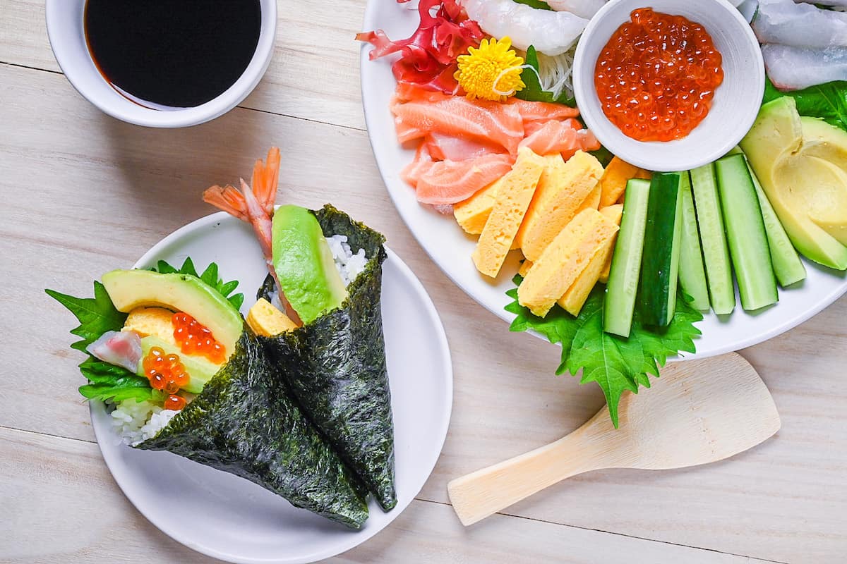 two temaki sushi hand rolls on a white plate with ingredients and soy sauce on the side
