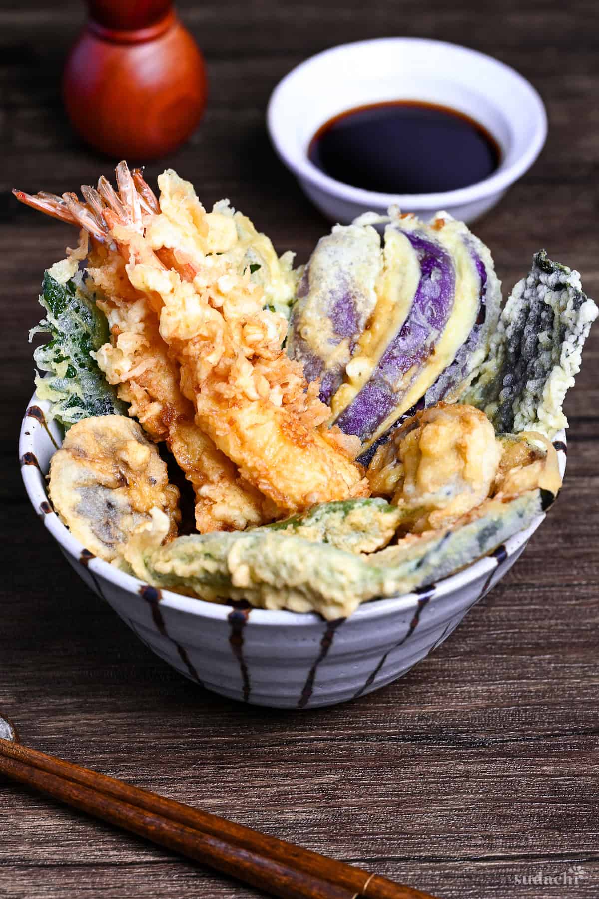 Tendon (Tempura Rice Bowl) made with shrimp and a variety of vegetables in a striped bowl on a wooden surface with homemade sauce in the background