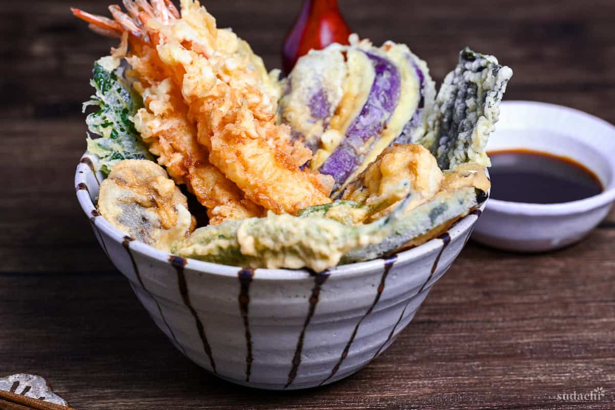 Tendon (Tempura Rice Bowl) made with shrimp and a variety of vegetables in a striped bowl on a wooden surface with homemade sauce in the background