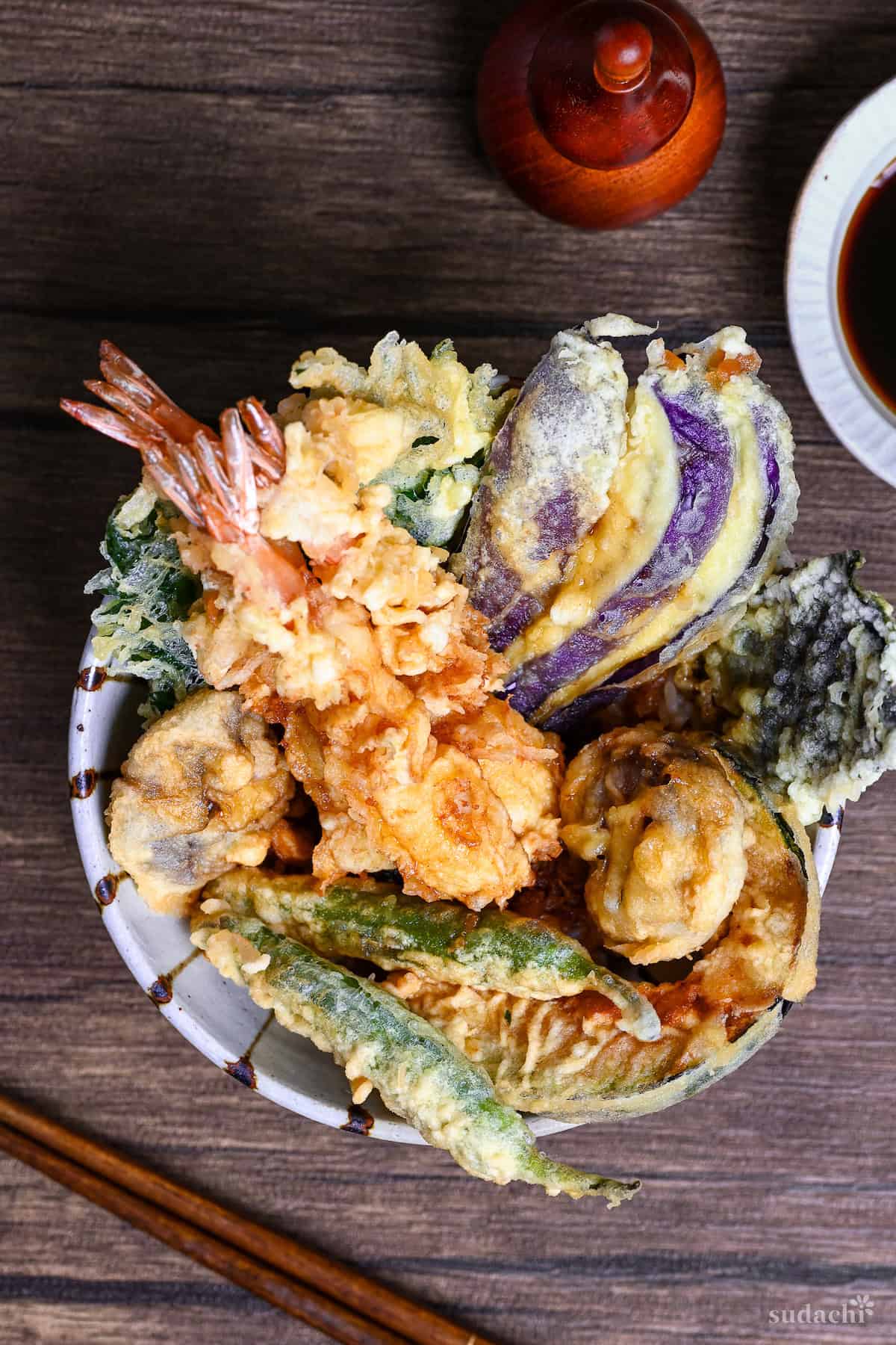 Tendon (Tempura Rice Bowl) made with shrimp and a variety of vegetables in a striped bowl on a wooden surface with homemade sauce in the background