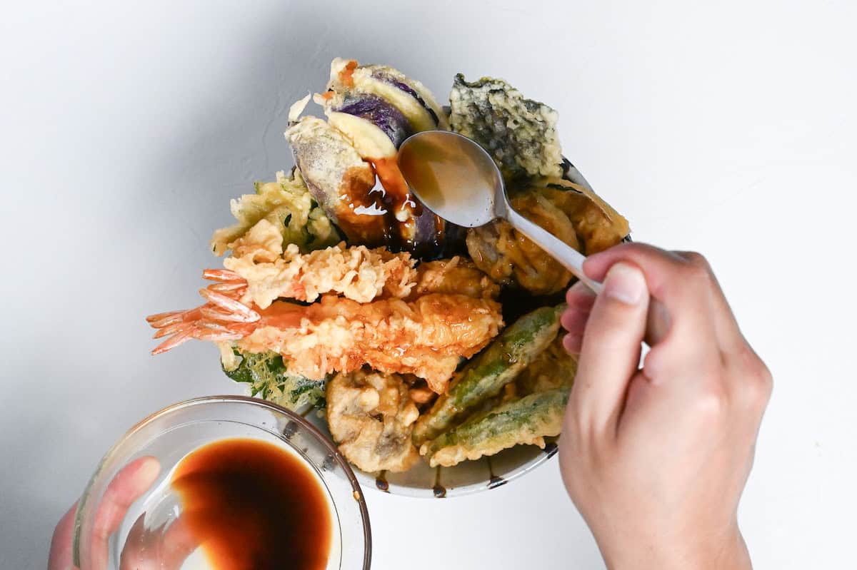 Tendon (Tempura Rice Bowl) made with shrimp and a variety of vegetables in a striped bowl drizzled with homemade sauce on a white background