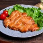 Sliced teriyaki chicken on a blue plate garnished with frilly lettuce, sliced cucumber and whole mini tomatoes