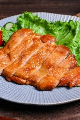 Sliced teriyaki chicken on a blue plate garnished with frilly lettuce, sliced cucumber and whole mini tomatoes