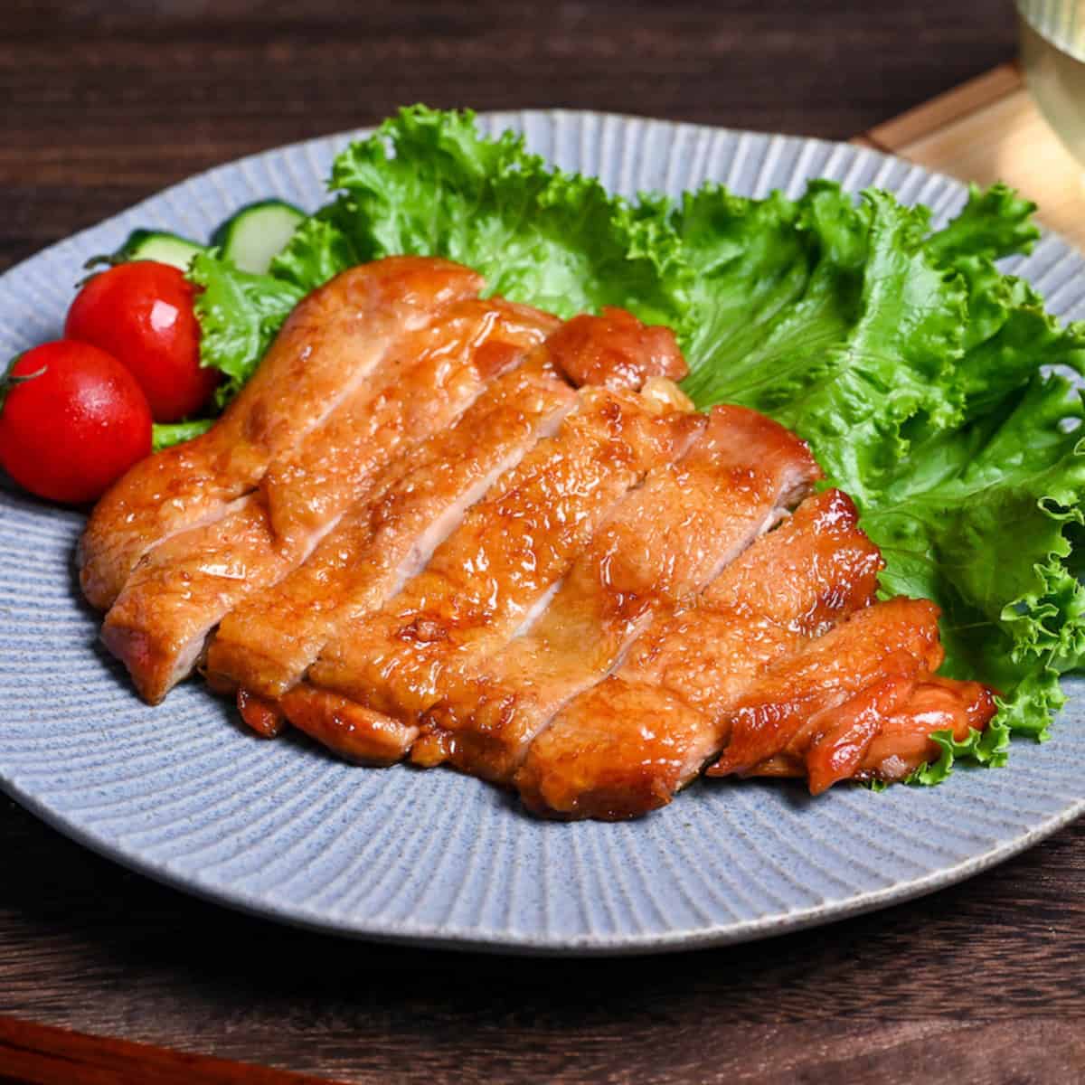 Sliced teriyaki chicken on a blue plate garnished with frilly lettuce, sliced cucumber and whole mini tomatoes