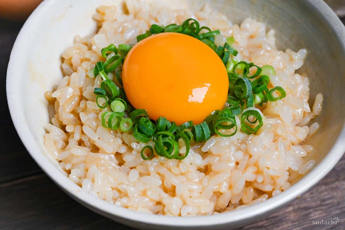 Tamago kake gohan topped with chopped green onions and a raw egg yolk in a cream ceramic bowl closeup