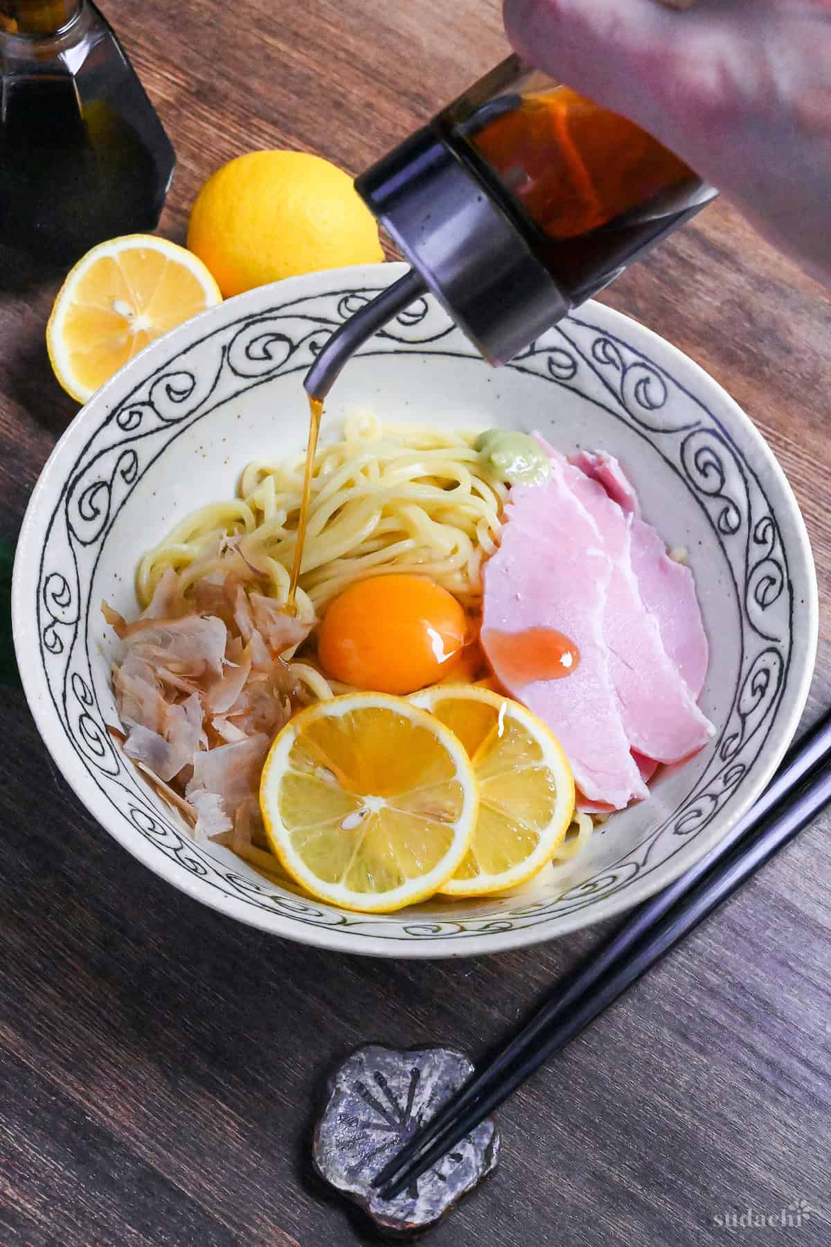 Pouring sesame oil over TKM topped with bonito flakes, a raw egg, ham, wasabi and lemon slices