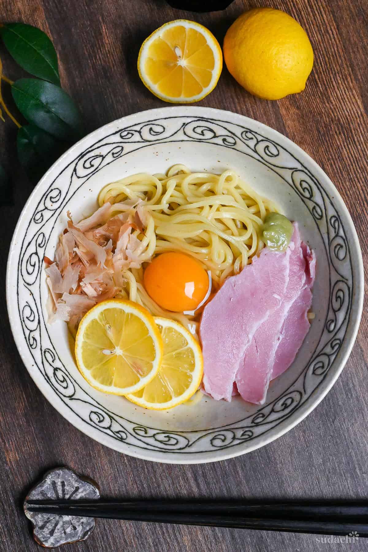 TKM (Tamago Kake Men) topped with bonito flakes, lemon slices, ham, wasabi and a raw egg served in a white bowl with black swirl pattern on rim on a dark wooden surface with lemon, soy sauce and leaves in the background top down