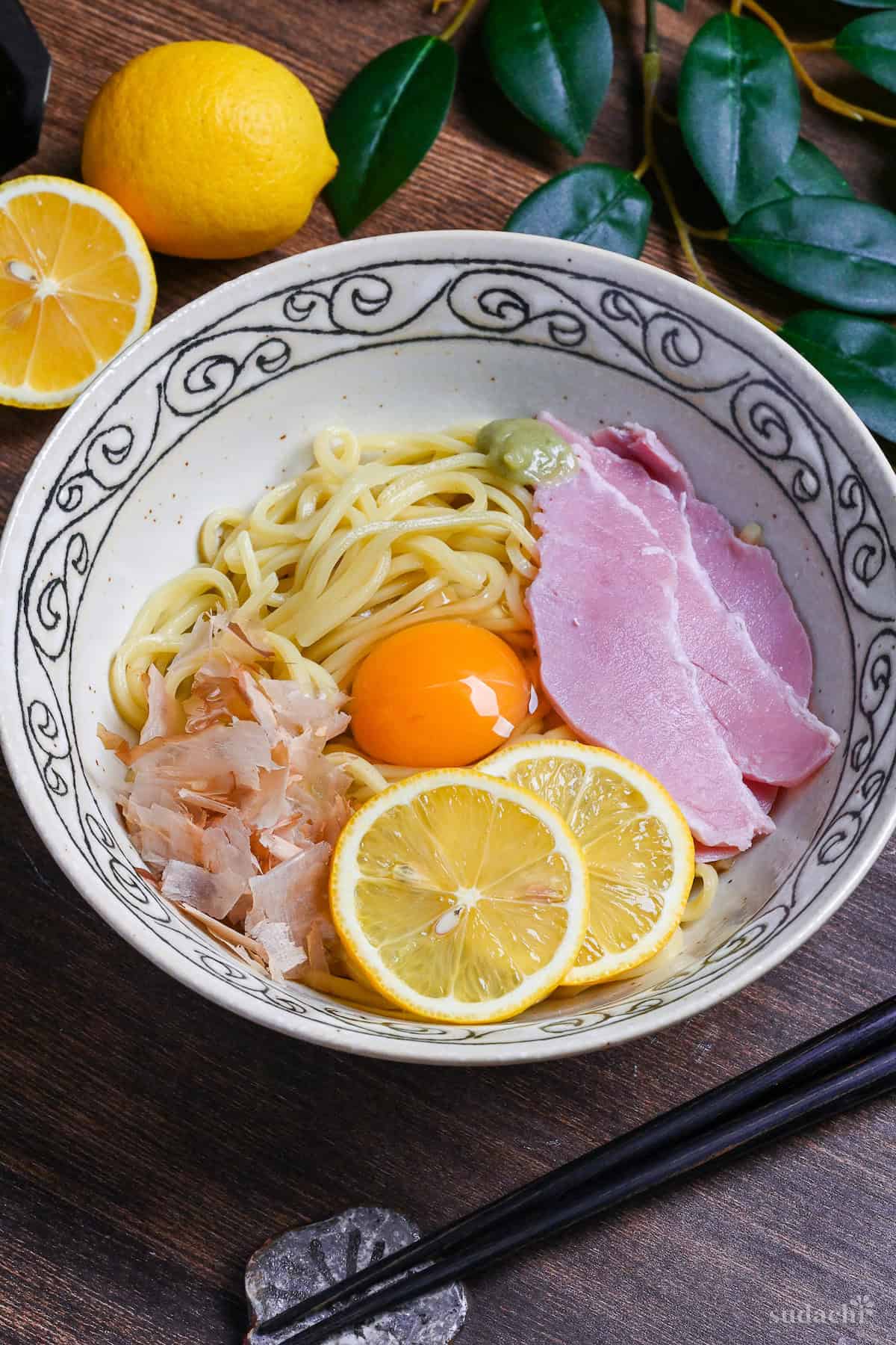 TKM (Tamago Kake Men) topped with bonito flakes, lemon slices, ham, wasabi and a raw egg served in a white bowl with black swirl pattern on rim on a dark wooden surface with lemon, soy sauce and leaves in the background
