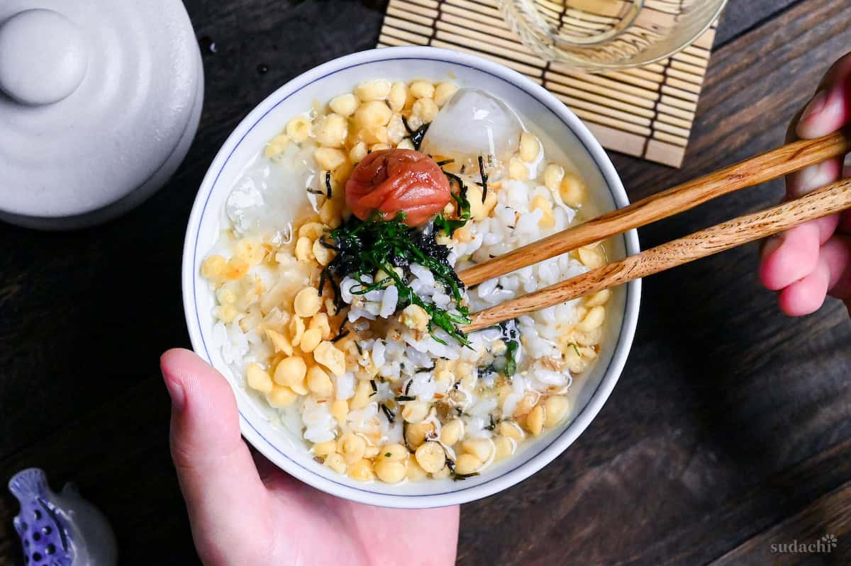 A hand holding cold plum chazuke in a white and blue bowl with wooden chopsticks