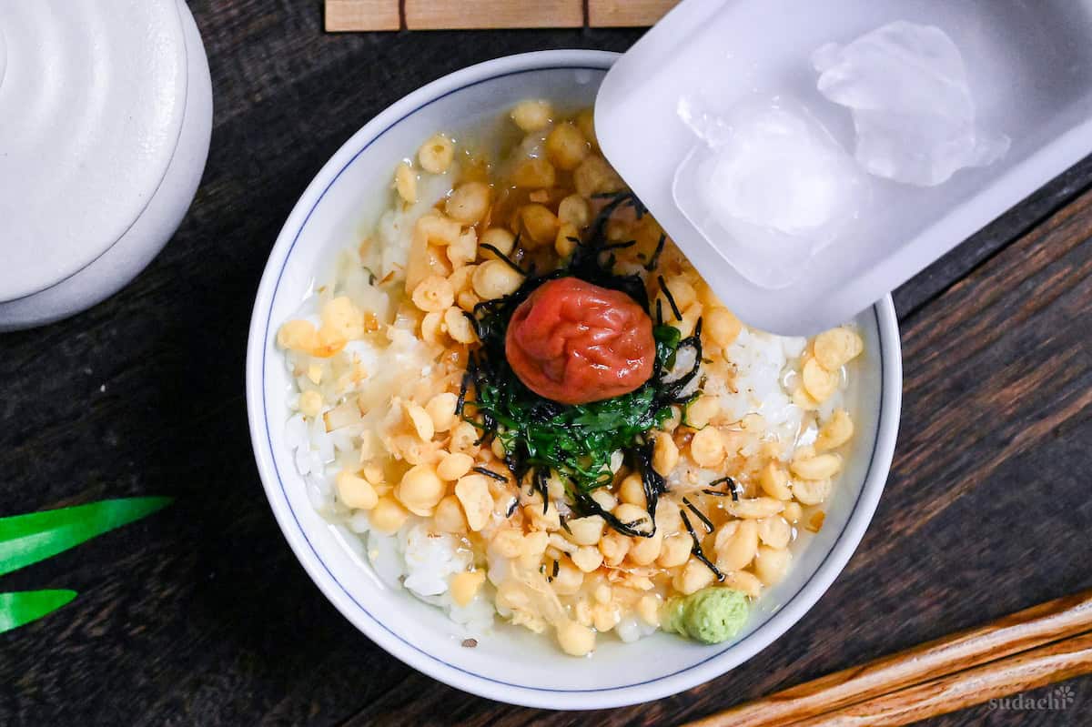 a white scoop with two ice cubes hovering above a bowl of hiyashi ume chazuke