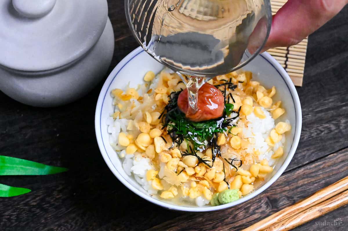 pouring chilled green tea from a glass into a bowl of rice with toppings and umeboshi pickled plum (ume chazuke)