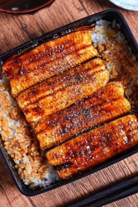 grilled eel coated with homemade unagi sauce and sprinkled with sansho pepper served over rice in a black lacquerware box (unaju) with a plate of pickles and bowl of clear soup