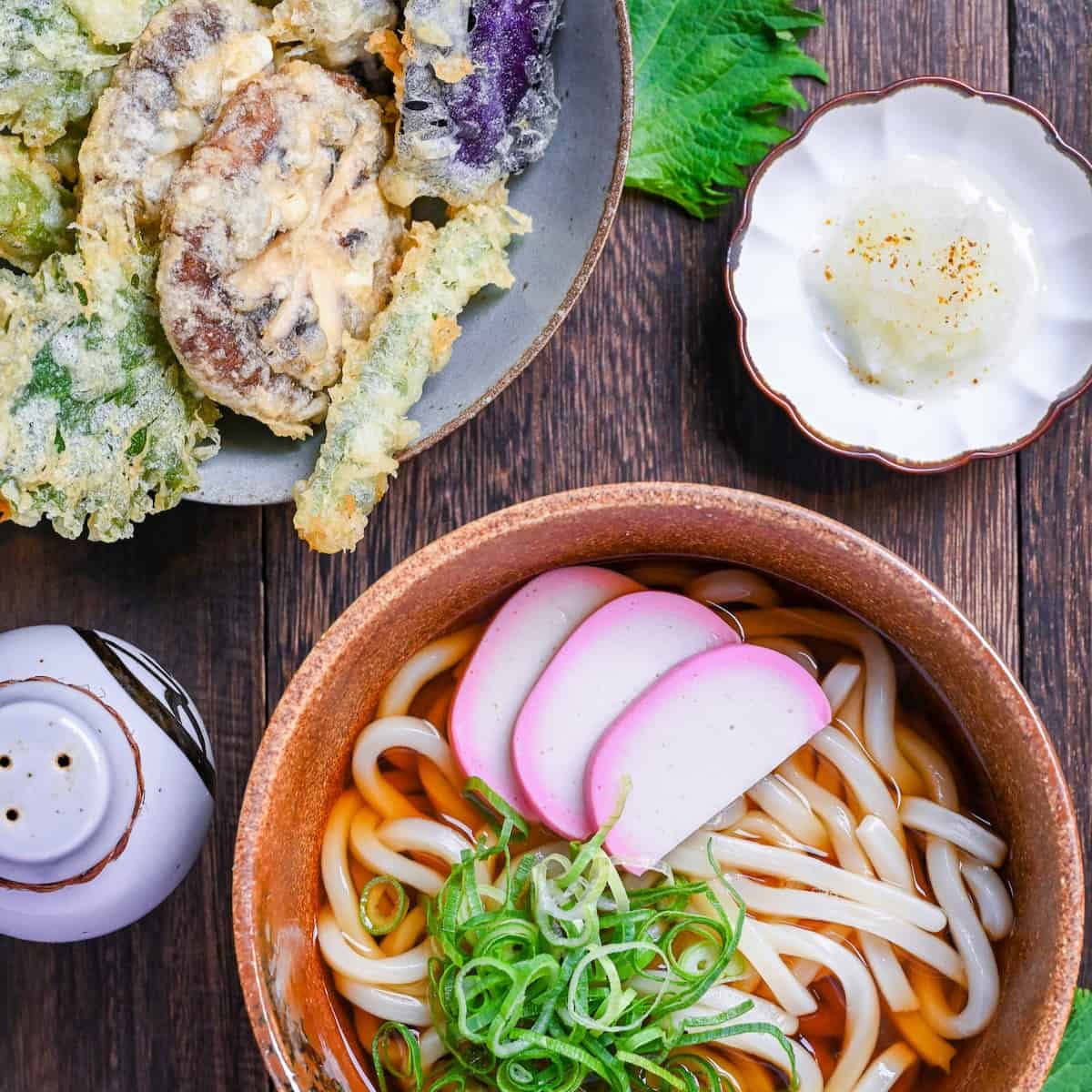 Udon noodles in simple broth in a brown bowl next to a variety of vegetable tempura including shiitake mushrooms, asparagus, shiso leaves and eggplant