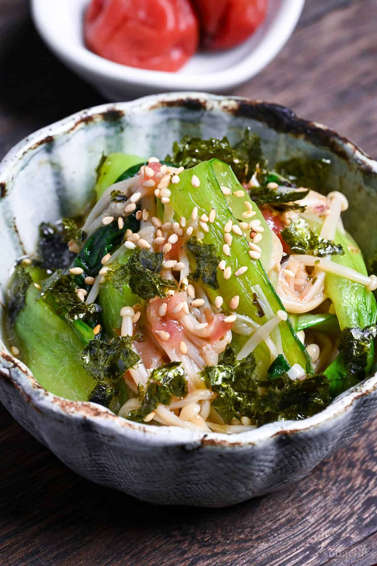 Wasabi-Ae salad with enoki mushrooms and bok choy in a blue and brown bowl on a dark wooden background with umeboshi on a white dish in the background closeup