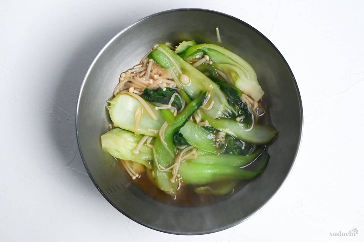 Bok choy and enoki mushrooms in broth in a steel mixing bowl on a white background