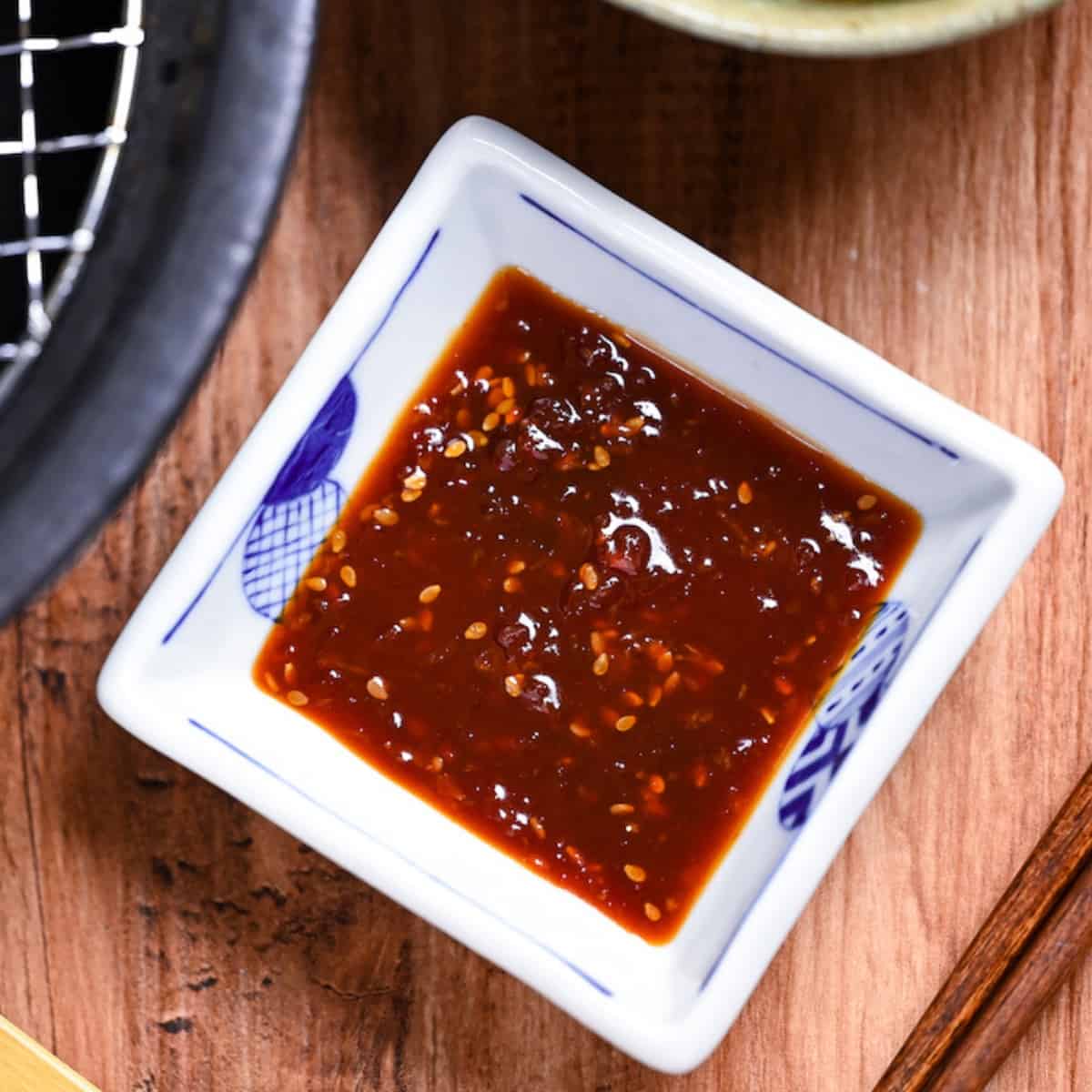 Homemade Japanese yakiniku sauce in a small square white bowl with blue border next to slices of thinly sliced beef on a metal grill
